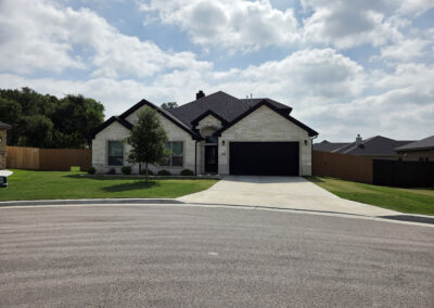 One-story tan brick home with gabled portico entry, two-car garage, and sidewalk in a quiet neighborhood