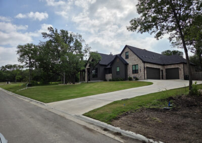Newly constructed dark-roofed home with mature trees and an extended concrete driveway leading to the garage