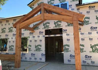 Residential entry porch under construction with a heavy timber gable over house-wrapped framing.