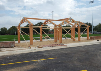 A row of repeating heavy timber king-post trusses forming an open-air covered structure under a clear sky.